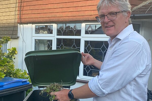 Edward Barnard in shirtsleeves place garden rubbish into a green waste wheelie bin