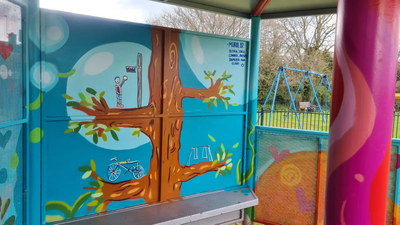 Photo of rain shelter for teenagers with brightly coloured decorative "graffiti" of a tree with bicycles and swings... in the background, the playground's swings can be seen